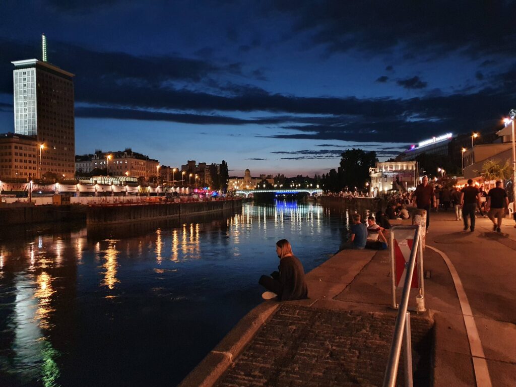 Ludzie siedzący na bulwarach nad Kanałem Dunajskim w Wiedniu nocą